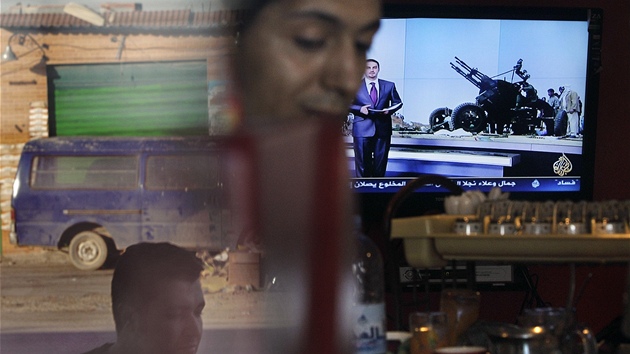 Majitel kavárny v Benghází pipravuje preso, zatímco televize za ním vysílá jednání v Dauhá, a v odrazu skla je vidt mu kráející ulicí (13. dubna 2011)