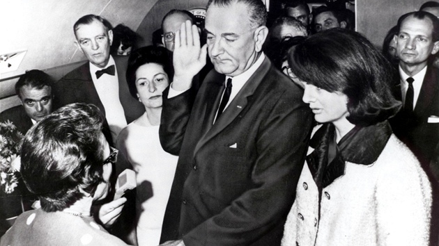 Lyndon Johnson skld prezidentskou psahu na palub letounu Air Force One. Vedle nj stoj Jackie Kennedyov, jet v obleen potsnnm krv prv zesnulho manela.