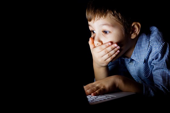 Young boy covered her mouth in fright, sitting at a computer on a dark night. The child is shocked by what he saw on the laptop.