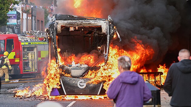 Lidé sledují příjezd hasičů k hořícímu vozidlu zapálenému během protestů ve východní části severoirského Belfastu. (9. června 2026)
