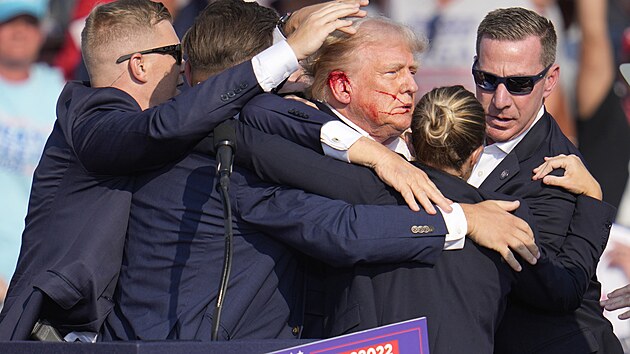 Donald Trump se sna zstat na nohou, obklopen agenty, kte ho rychle evakuuj ze scny po pokusu o atentt. Krev mu stk po tvi.