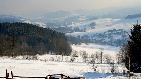 Výhled do údolí s rozptýlenou zástavbou obce Telecí, na pozadí se v oparu ztrácí členité panorama Žďárských vrchů