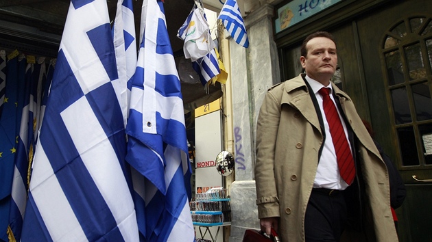 Mu jde po ulici kolem kiosku prodávajícího ecké vlajky.