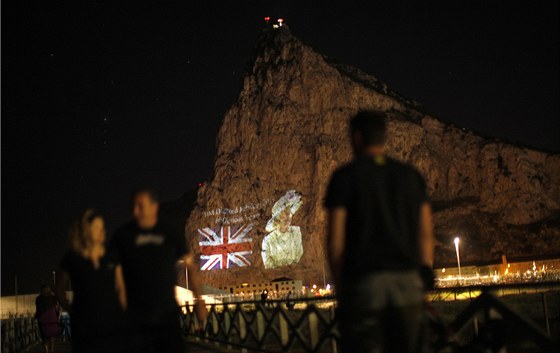 Lidé sledují ze panlských beh obrázek britské královny Albty II. a vlajky