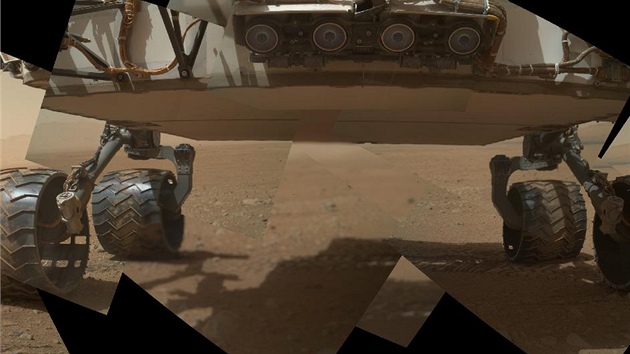 Prvn zbr na podvozek Curiosity na Marsu z 9. z 2012. Nahoe je tveice kamer, kter maj dvat pozor na pekky. Na kolech je u vidt usazen jemnozrnn maransk prach. Spodek voztka je asi 66 cm nad zem. Na obzoru vpravo je st hory s tmavmi dunami na pat. Voztko zatm ujelo nco mlo pes sto metr.