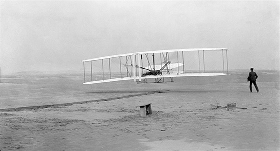 První let motorového letadla v historii. Řídí Orville Wrightr, po straně beží...