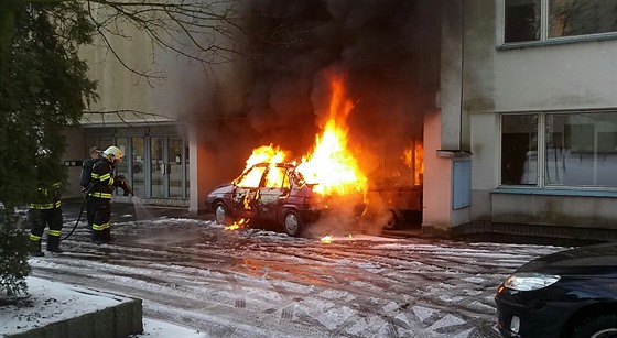 Hoící automobil v sousedství domu, kterému plameny pokodily fasádu.