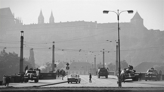 Pochmurn� odpoledne na M�nesov� most� kontrolovan�m sov�tsk�mi voj�ky po srpnov� invazi. Naho�e vystupuje nezam�niteln� silueta ��sti Pra�sk�ho hradu, dvojice �t�hl�ch v�� pat�� bazilice svat�ho Ji��, zcela vpravo je �ern� v�. Z techniky vid�me t�i kusy sov�tsk�ch kolov�ch obrn�n�ch transport�r� BTR-152 a tank typov� �ady T-54/55. P�es most p�ejela �Carevna", neboli Volha GAZ-21, tuzemsk�ho u�ivatele. (Srpen 1968, Praha)