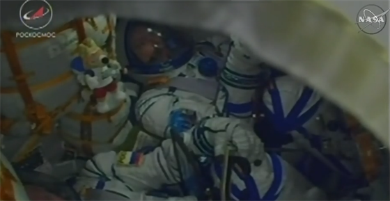 Posádka kosmické lodi Sojuz MS-08 bhem startu 21.3.2018. Krteka pipevnného k okraji kesla zakrývá výrazn vtí maskot výpravy.