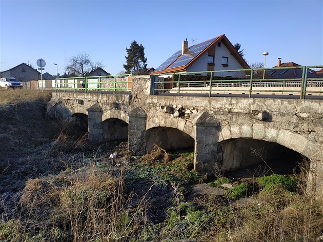 Pvodní kamenný most pes Pilský potok pekrývá asfaltová silnice s kovovým...