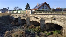 Pvodní kamenný most pes Pilský potok pekrývá asfaltová silnice s kovovým...
