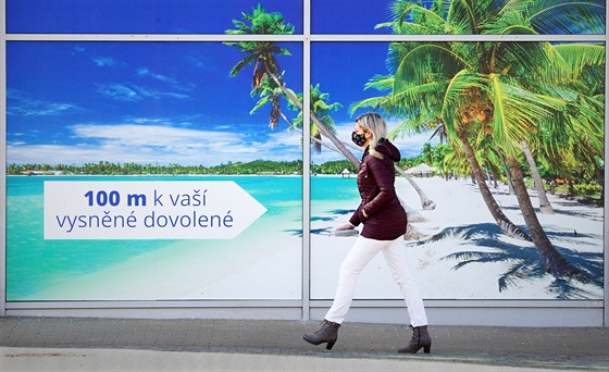 Žena v roušce prochází podél reklamního poutače cestovní kanceláře