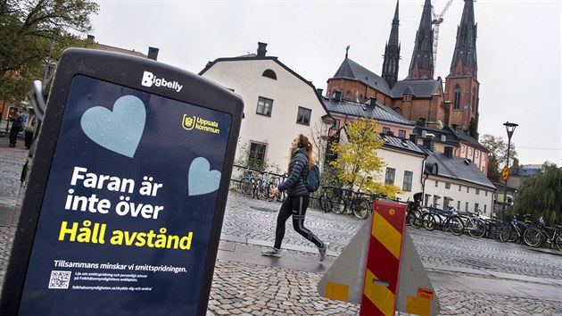 Lidé procházejí kolem koše s nápisem „Nebezpečí neskončilo - udržujte si odstup“ na pěší zóně v centru Uppsaly ve Švédsku. (21. října 2020)