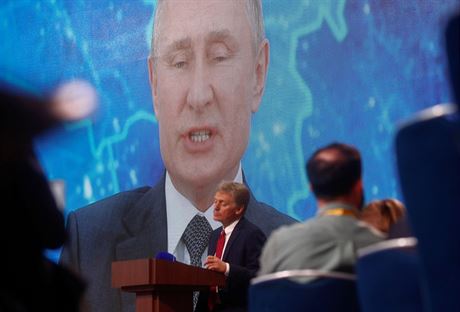 Mluví Kremlu Dmitrij Peskov stojí u pultu, zatímco z plátna za jeho zády promlouvá jeho éf, Vladimir Putin