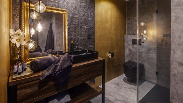 Luxusní koupel. Kontrastem hladkému šedému základu koupelny jsou reliéfní obklady z kolekce Aparici, zlaté povrchy, kresba dřevěného nábytku a netradiční černá matná sanita.