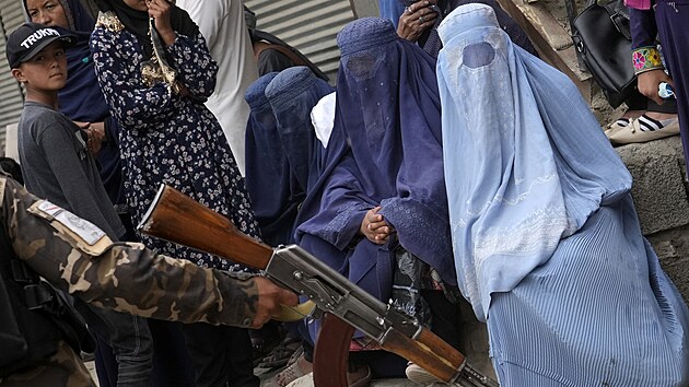 Bojovník Tálibánu drží zbraň a stojí na stráži, zatímco ženy čekají na potravinové příděly. (18. května 2022)