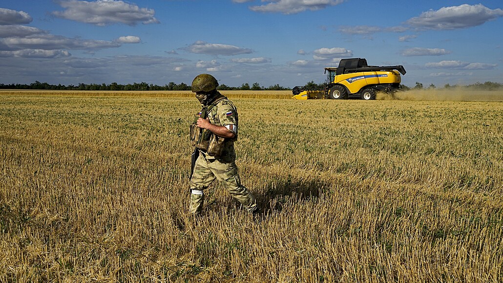 Ruský voják steí oblast, zatímco farmái sklízí penici na poli nedaleko...