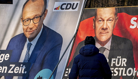 Mu prochází kolem volebních plakát, na kterých je vyobrazen nmecký kanclé...