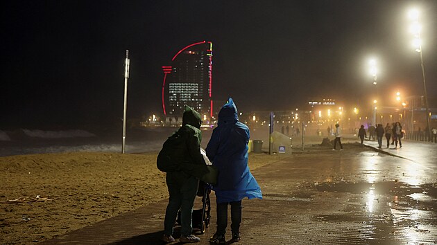 P�r se proch�z� po pl�i La Barceloneta, zat�mco za nimi v Barcelon� nar�ej� velk� vlny z p�ech�zej�c� bou�e. (26. prosince 2025)